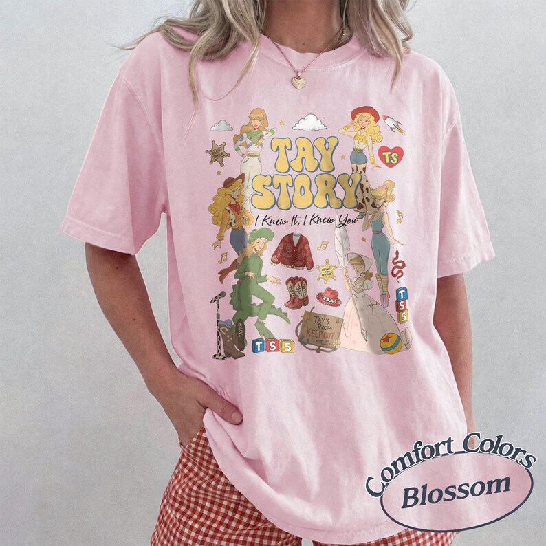 May include: A light pink short-sleeve t-shirt featuring a graphic print with the text TAY STORY I Knew It, I Knew You. The design includes various illustrated figures, a red cardigan, a cowboy boot, a hat, and a sign reading TAY'S ROOM KEEP OUT.
