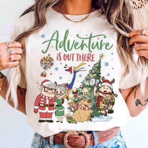 May include: Cream-colored t-shirt featuring a colorful graphic with the text "Adventure is Out There." The design includes cartoon characters, a Christmas tree, a hot air balloon, and snowflakes, creating a festive and adventurous theme.