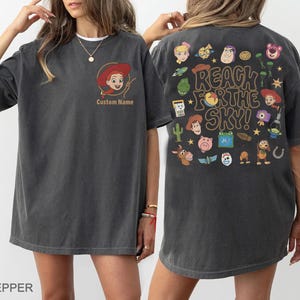May include: A charcoal gray t-shirt featuring a small graphic of Jessie on the front with the text Custom Name. The back displays a large collage of various Toy Story character illustrations and the text REACH FOR THE SKY in a bold font.