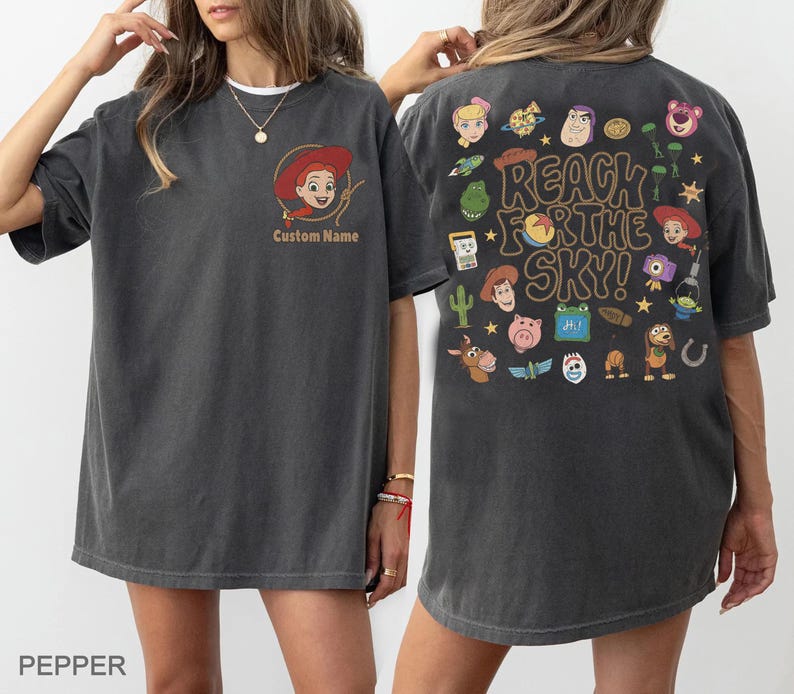 May include: A charcoal gray t-shirt featuring a small graphic of Jessie on the front with the text Custom Name. The back displays a large collage of various Toy Story character illustrations and the text REACH FOR THE SKY in a bold font.