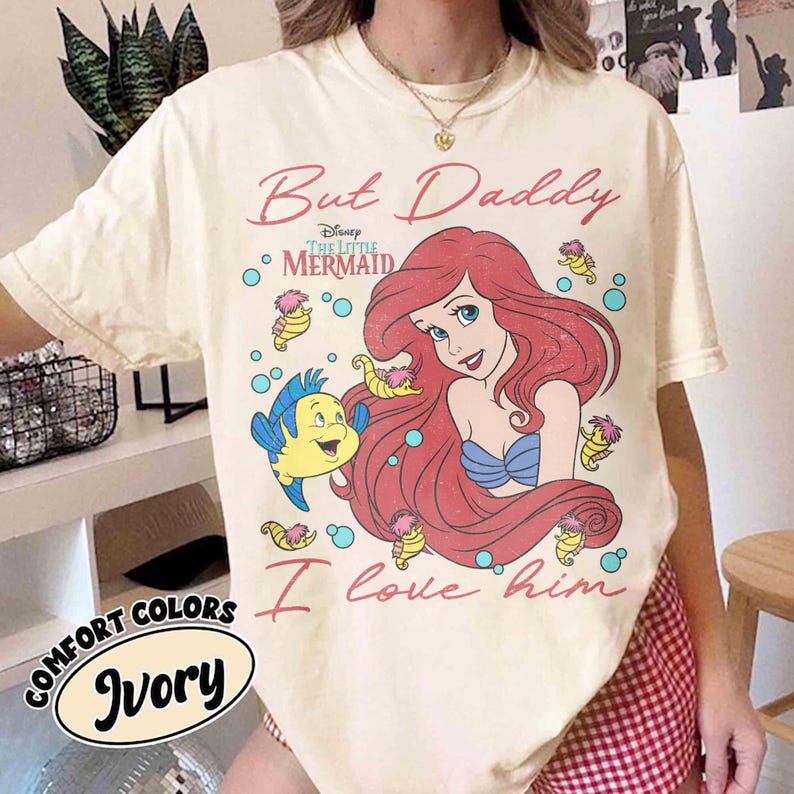 May include: An ivory t-shirt featuring a graphic print of Ariel from The Little Mermaid and Flounder, surrounded by small seahorses and bubbles. The text reads But Daddy I love him and includes the Disney The Little Mermaid logo in red and blue.