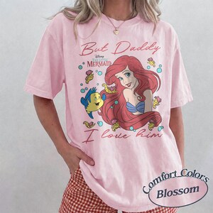 May include: A light pink short-sleeve t-shirt featuring a graphic of Ariel and Flounder from The Little Mermaid. The text reads But Daddy I love him. The design includes small seahorses and bubbles in blue and yellow tones surrounding the characters.