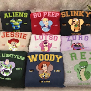Puede incluir: Una colección de sudaderas coloridas con personajes de la película animada Toy Story. Cada sudadera muestra el nombre de un personaje en letras negritas, junto con una imagen del personaje y el texto "EST 1995".