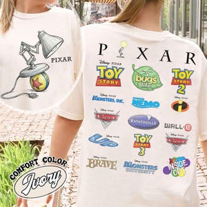 Może przedstawiać: Kremowa koszulka z ilustracją lampy Pixar i napisem "PIXAR". Z tyłu znajdują się loga filmów Pixara, takich jak "Toy Story", "Dawno temu w trawie" i "Gdzie jest Nemo". Koszulka jest oznaczona jako "Comfort Color".