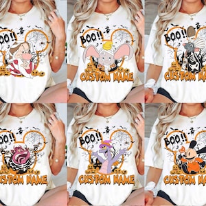 May include: A white t-shirt featuring a Halloween-themed graphic with a mouse-ear silhouette, spider webs, pumpkins, and the text "BOO!!" and "CUSTOM NAME". The design includes various cartoon characters like Dumbo, Remy, and others.