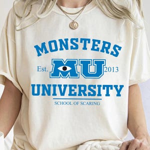 May include: Cream-colored t-shirt featuring the text "MONSTERS UNIVERSITY" in blue, with "MU" in a stylized logo and the year "2013". Below, it reads "SCHOOL OF SCARING". A gold necklace is visible.