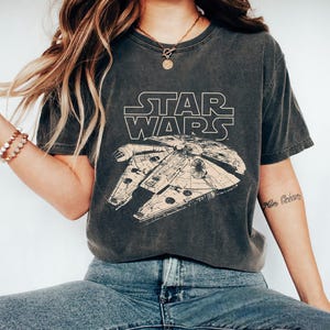 Può includere: T-shirt grigio scuro con il logo di Star Wars e un'illustrazione dettagliata del Millennium Falcon in beige chiaro. La maglietta ha maniche corte e girocollo.