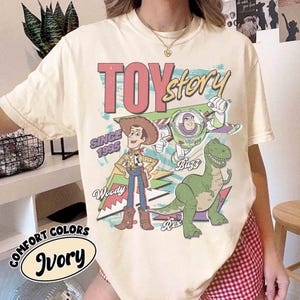 May include: Ivory-colored t-shirt featuring the words "Toy Story" in a retro font, with Woody, Buzz Lightyear, and Rex the dinosaur. The shirt also includes the text "Since 1995" and "Comfort Colors Ivory".