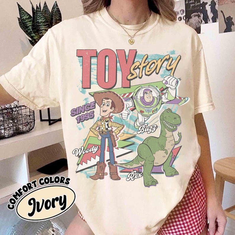 May include: Ivory-colored t-shirt featuring the words "Toy Story" in a retro font, with Woody, Buzz Lightyear, and Rex the dinosaur. The shirt also includes the text "Since 1995" and "Comfort Colors Ivory".
