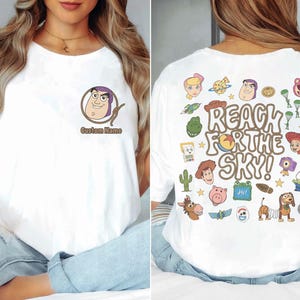 May include: A white t-shirt featuring a small Buzz Lightyear graphic on the front chest with the text Custom Name. The back displays a large colorful collage of Toy Story characters and the text REACH FOR THE SKY in a brown bubble font.