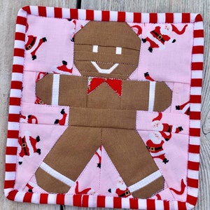 Peut inclure: Un dessous de plat carré et matelassé de Noël avec un motif de bonhomme en pain d'épices. Le bonhomme en pain d'épices est marron avec des accents blancs et un nœud papillon rouge. Le fond est rose avec un motif de Père Noël, et la bordure est à rayures rouges et blanches.