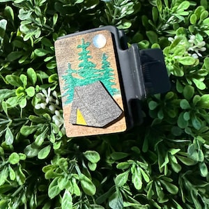May include: A black phone grip with a wooden panel featuring a camping scene. The panel shows a gray tent with a yellow corner, green trees, and a white moon against a brown background.