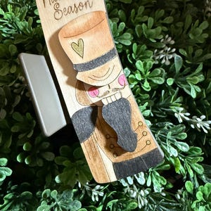May include: Wooden bookmark featuring a Nutcracker soldier design. The bookmark has the words "Nutcracker Season" at the top. The Nutcracker has a gold hat, pink cheeks, and a black uniform. The bookmark is set against a backdrop of green foliage.