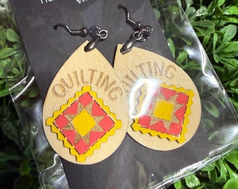 May include: Teardrop-shaped wooden earrings featuring the word QUILTING engraved at the top and a central square quilt block design in red, yellow, and orange. The earrings hang from dark metal hooks and are attached to a black display card.