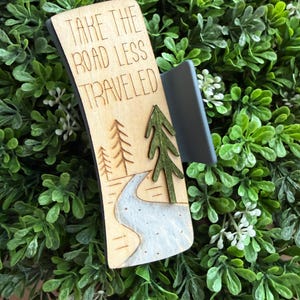 May include: Wooden sign with the text "TAKE THE ROAD LESS TRAVELED". The sign features a laser-cut design of a road, trees, and a river. The sign is attached to a gray clip.