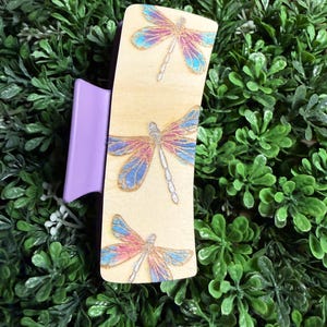May include: A light-colored wooden hair clip with three dragonfly designs. Each dragonfly has pink, blue, and silver accents. The clip itself is a solid lavender color. The background is a bed of green leaves.