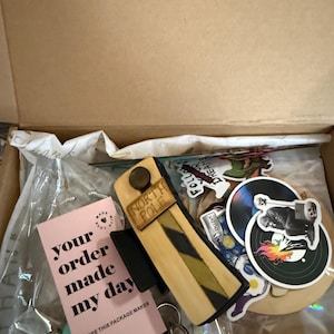 May include: A cardboard shipping box containing a variety of items including a wooden North Pole sign, a pink card with the text your order made my day, several circular stickers featuring graphic designs, and a silver metal keychain with beads.