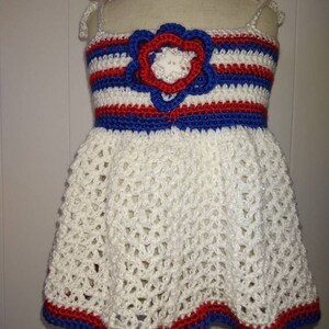 May include: A white crocheted dress with red, white, and blue stripes and a crocheted flower on the bodice. The dress has a scalloped hem.