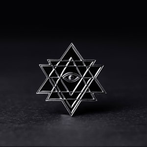 May include: A silver-colored metal pin with a geometric design featuring a star with an eye in the center. The pin is on a black background.