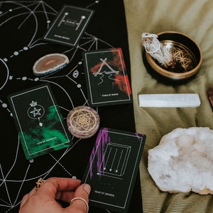 May include: Tarot cards spread out on a black cloth with a white geometric design. The cards include "King of Pentacles", "Three of Wands", and "Four of Swords".