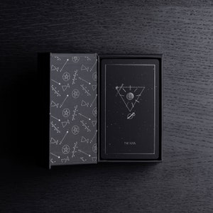May include: A black tarot card deck box with a white geometric pattern on the side. The box is open, revealing a black tarot card with a white illustration of a figure standing on a cliff, with a star above their head. The card reads "The Fool".