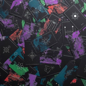 May include: A deck of tarot cards with black backgrounds and white geometric designs. Each card has a different design and is painted with a variety of colors, including purple, green, and blue.