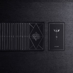May include: A black tarot deck with silver foil accents. The card face up is The Lovers card, which features a heart with wings and a star.