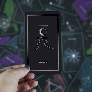 May include: A black tarot card with a white crescent moon and a winding path design. The card reads "THE MOON" at the bottom.