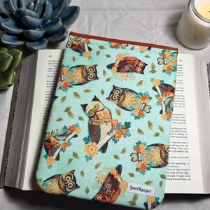 May include: A teal blue book cover with a repeating pattern of owls and flowers. The owls are brown, orange, and yellow with intricate details. The text "StoryKeeper" is printed on the bottom right corner of the cover.