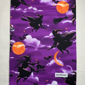 May include: Purple fabric with a repeating pattern of black silhouettes of witches flying on broomsticks with orange moons and stars. The fabric has the text "StoriKeeper" in the bottom right corner.