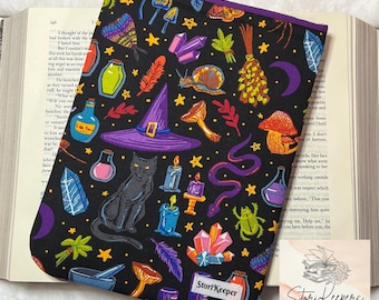 May include: Black book sleeve with a purple border, featuring a whimsical witchy design. The pattern includes a black cat, witch hat, crystals, mushrooms, stars, and potion bottles. The text "StoriKeeper" is visible.
