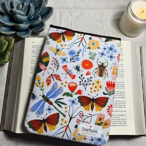 May include: A white book cover with a colorful floral and insect pattern. The cover features butterflies, dragonflies, beetles, and flowers in shades of red, yellow, blue, and green. The text "Story Keeper Handmade" is printed in the lower right corner.