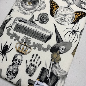 May include: A white fabric background with a black and white print of various Halloween themed images. The images include a skull, a spider, a witch, a coffin, a crown, a handprint, a butterfly, a clock, a rose, an eye, and a raven. The fabric has the text "StoriKeeper" printed on it.