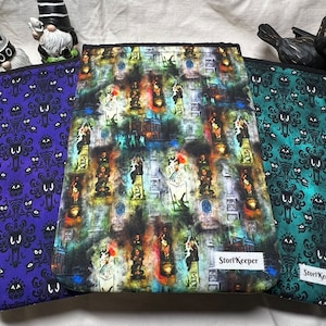 May include: Three different fabric zippered pouches with a Halloween theme. The pouches are purple, green, and black with a repeating pattern of ghosts. The black pouch has a colorful print of a haunted house with ghosts and other spooky characters. The pouches have a label that says "StoryKeeper".