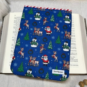May include: A blue book sleeve with a red and white striped top edge. The sleeve features a repeating pattern of snowmen, reindeer, and Santa Claus on a blue background. The text "StoryKeeper" is printed on the bottom right corner of the sleeve.