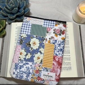 May include: A patchwork fabric book sleeve with a floral design. The sleeve has a blue, green, yellow, pink, and white color scheme. The sleeve has a tag that says "Story Keeper Handmade".