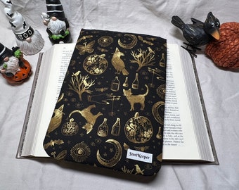 May include: A black book cover with a gold foil design featuring a cat, a crow, a pumpkin, and other Halloween-themed imagery. The text "StoryKeeper" is printed in gold foil on the bottom of the cover.