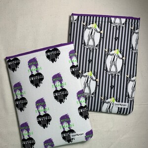 May include: Two fabric cases with purple trim. One case has a white background with a repeating pattern of a character's face and the words "MYSELF STRANGE UNUSUAL". The other case has a black and white striped background with a repeating character design.
