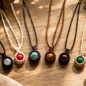 May include: Six necklaces featuring round gemstones in macrame settings. The stones include a translucent white, bright red, turquoise, tiger eye, dark red, and green malachite. Each pendant hangs from a matching braided cord in black, cream, or brown.
