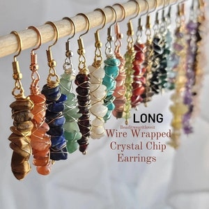 Puede incluir: Una colección de pendientes largos de cristal envueltos en alambre. Los pendientes están hechos con varios tipos de cristales, incluyendo turquesa, amatista y cornalina. El alambre es de color dorado y los pendientes cuelgan de una pantalla de madera.