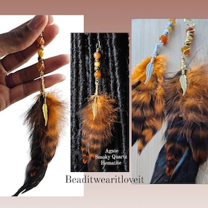 以下が含まれることがあります： 羽根とビーズが付いたヘアタイが2つ。ビーズはアゲート、スモーキー・クォーツ、ヘマタイト。羽根は茶色と黒で、金色のアクセントが付けられている。"Agate Smoky Quartz Hematite Beaditwearitloveit"というテキストが見える。