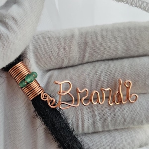 Op de afbeelding: Een haaraccessoire van koperdraad met de naam Brandi in cursief schrift, bevestigd aan een spiraal van koperdraad met twee kleine groene kralen, gewikkeld rond een enkele dikke lok zwart haar.