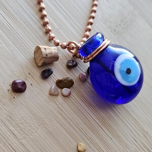 May include: A blue glass evil eye pendant necklace with a copper-colored chain. The pendant has a white and blue eye design. A cork and small stones are next to the necklace on a wooden surface.