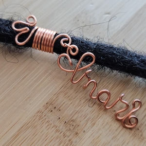 Op de afbeelding: Koperdraad haarjuwelen met het woord "Share" op een zwarte dreadlock. De sieraden bevatten een gewikkelde spiraal en decoratieve krullen. De achtergrond is een lichtgekleurd houten oppervlak.