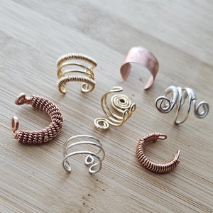 May include: A collection of seven ear cuffs made of wire in various colors and styles. The ear cuffs are arranged on a wooden surface.