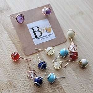 May include: Assortment of wire wrapped gemstone earrings. The earrings feature various stones including amethyst, turquoise, lapis lazuli, carnelian, and clear quartz. The earrings are displayed on a wooden surface with a small card that reads "Bead It Wear It Love It Handmade Jewelry".