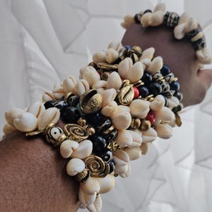 May include: A multi-layered bracelet set featuring numerous white cowrie shells, smooth black beads, and small metallic gold-toned charms with spiral and circular patterns. The bracelets are worn on the wrist, showcasing a dense, textured arrangement.