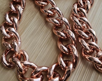 May include: A rose gold chain necklace with large, interlocking links. The chain is displayed on a wooden surface, with a decorative wooden tray in the background. The necklace is a statement piece.