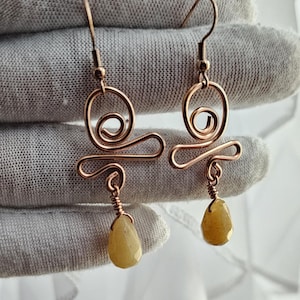 Op de afbeelding: Een paar handgemaakte koperdraad oorbellen. Elke oorbel heeft een wervelend ontwerp met een druppelvormige, gele edelsteen hanger. De oorbellen worden gepresenteerd tegen een zachte, neutrale achtergrond.