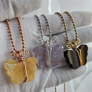 May include: Three butterfly pendant necklaces. The pendants are made of gemstones in yellow, purple, and brown. Each butterfly is wire-wrapped and hangs from a ball chain necklace in copper, silver, and gold colors.